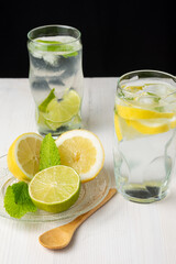 Top view of two glasses of water with lemon and lime slices with ice cubes on white wood, with wooden spoon, plate with half lemons and mint, on black background, vertical
