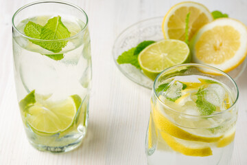 Top view of two glasses of water with lemon and lime slices with ice cubes on white wood, plate with half lemons and mint, vertical