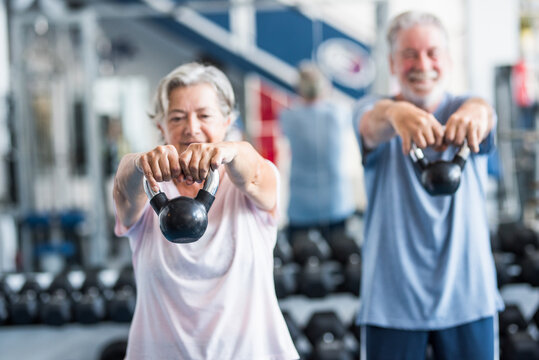 couple of two active and healthy seniors or pensioners or mature people doinng exercise together in the gym holding a dumbbell in their hands and doing squats - fitness lifestyle dieting concept