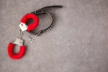 Black leather lash and metal handcuffs with red fur on a gray background, top view