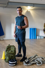 Portrait of motivated boot camp instructor in gym hall. Rope, dumbbells and sandbag on wooden floor.