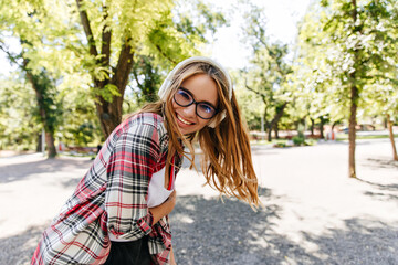 Glad long-haired girl enjoying music in park. Glamorous blonde lady in headphones dancing in sunny day.