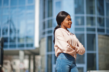Fototapeta premium Hipster african american girl wearing pink hoodie, jeans posing at street against office building with blue windows.