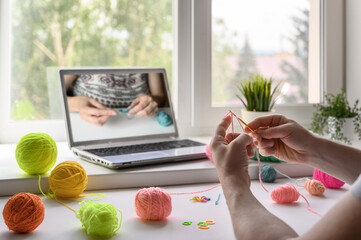 Selective focus on the hands of a man sitting at the table and watching a video step by step guide on crocheting. He holds a hook and yarn in his hands.
