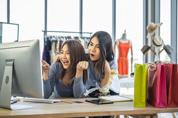 Two young Asian girls shop owners at work desk inside a clothing store looking and pointing at computer monitor with happy cheerful wow surprised expression.