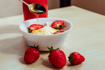 cottage cheese in a white bowl with sour cream and honey pouring from a spoon. Strawberries lying nearby. healthy breakfast.