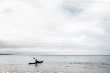 A man using a blue Kayak in the Ocean on a still but cloudy day