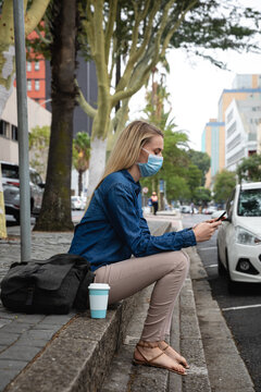Caucasian woman wearing a covid19 coronavirus mask sitting on a sidewalk and using her phone