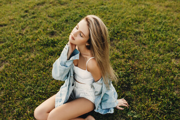 Young attractive woman with long blonde hair, with eyes closed, wearing a denim jacket, sitting on green grass.