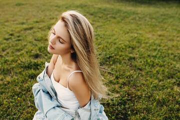 Fototapeta na wymiar Young attractive woman with long blonde hair, with wind in hair, wearing a denim jacket, sitting on green grass.