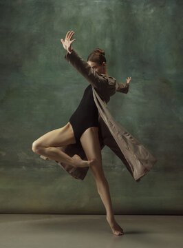 Warm. Graceful classic ballerina dancing, posing isolated on dark studio background. Stylish trench coat. Grace, movement, action and motion concept. Looks weightless, flexible. Fashionable, style.