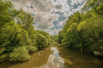 Fototapeta na wymiar view from a bridge on calm river surrounded by green trees and mirroring blue sky with clouds