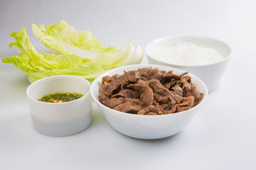 isolated close up shot of a bowl of sliced beef fillet pieces, a plate of cellophane glass noodles, a cup of Vietnamese Nuoc Cham sauce and a pile of Chinese napa cabbage leaves on a white background