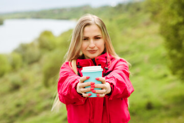 Cup of hot beverage, coffee to go cup in the woman's hands. Nature background. Spending time outdoors in summer,autumn, spring. Blonde woman is wearing pink jacket.