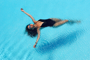 Young beautiful sexy slim tanned caucasian woman with brunette hair in black swimwear relaxing and lying in blue water of swimming pool at tropical resort. Vacation concept. Top poinit of view