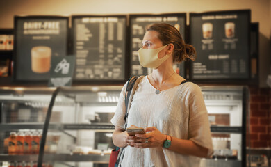 A young woman in a protective mask stands in a cafe at the counter with desserts and a cash register with a smartphone in her hands. New normal lifestyle