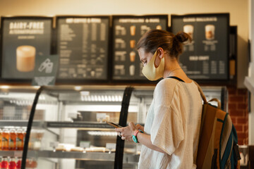 A young woman in a protective mask stands in a cafe at the counter with desserts and a cash register with a smartphone in her hands. New normal lifestyle