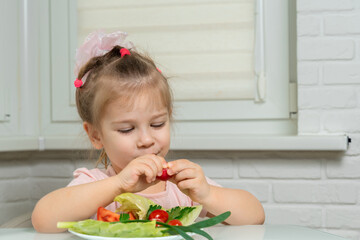 little girl sits at a table in the kitchen and eats vegetables