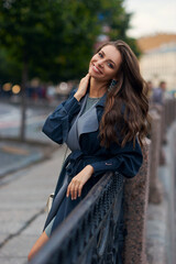 Fototapeta premium Happy joyous young woman standing and posing at river embankment in city. Pretty girl in dress and raincoat with makeup and wavy hair. Brunette caucasian female.