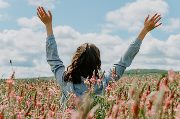 Back view of young woman in pink flower field with arms up towards the blue sky