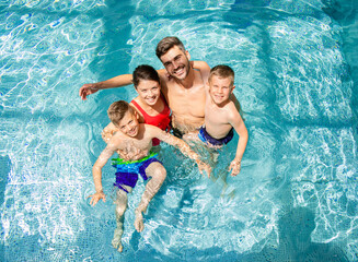 Top view of smiling family of four having fun and relaxing in indoor swimming pool at hotel resort. - obrazy, fototapety, plakaty