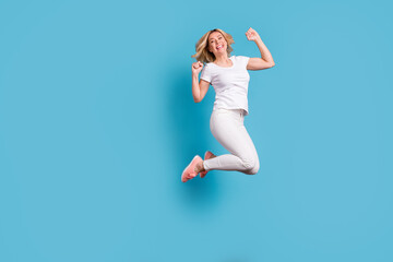 Fototapeta na wymiar Full body photo of attractive funny crazy lady jump high up celebrating holiday raise fists astonished excited wear casual white s-shirt pants footwear isolated blue color background