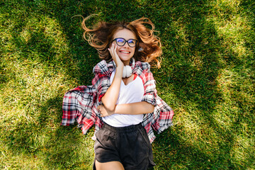 Dreamy girl in headphones lying on grass and looking at sky. Positive smiling woman spending weekend morning in park.