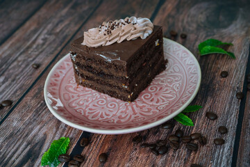 Piece of Chocolate cake with cream on plate. Chocolate cake on wooden background with coffee beans and mint leaves. Delicious slice of layered tart on plate. Baking and decorating chocolate cake
