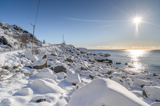 Sunrise from the sea in snowy road along the shore. Rocks are covered with white fluffy snow in the beautiful winter while the blight red orange sun rising from the horizon under clear blue sky. 