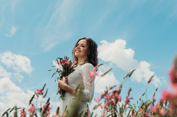 Mid body portrait of a young woman in white dress smiling to the side and holding a flower bouquet,...