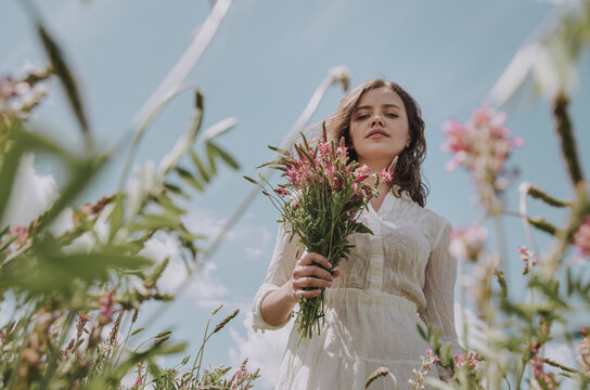 Low angle portrait of young woman with flower bouquet framed by growing flowers in a field