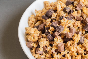 Granola crunchy muesly with pieces of dark and milk chocolate in a bowl on light brown background....