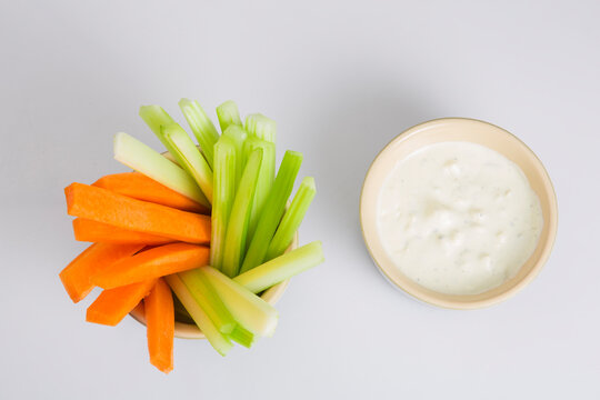 close up isolated flat lay top view shot of party snack food. A bowl of crunchy orange carrot and juicy green celery sticks with a white cup of blue cheese dipping sauce on a white background