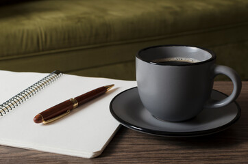 Closeup of cup of coffee, blank notebook and pen on the table against green sofa