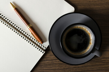 Top view of grey cup of coffee on the saucer, blank notebook and pen on the wooden table