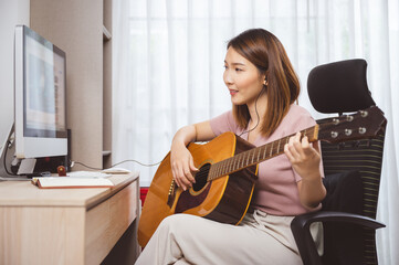 Asian woman guitar player and vlogger playing guitar in front computer at home while social...