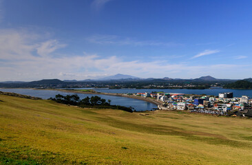 Fototapeta na wymiar awesome view of seaside small city within seongsun ilchubong jeju island south korea with yellowish grass foreground and slightly cloudy blue sky and ocean. Visible at far is mountain area