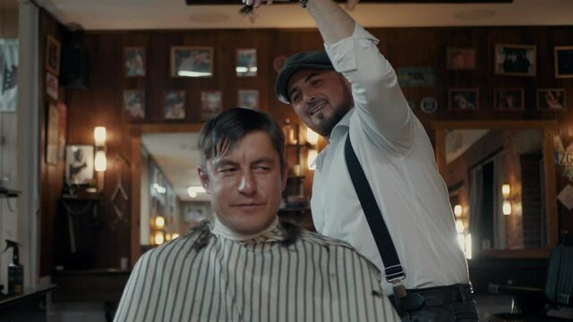 Barber mimics "Salt Bae" while cutting a client. Stylishly dressed Barber with a beard and a cap. Barber with a shirt and pants with suspenders. Parody of "Salt Bae" in the barbershop.