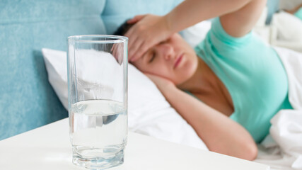 Glass of water on the bedside table against sick woman with migraine lying in bed