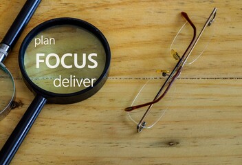 Conceptual image of plan focus deliver result in business. White text. Magnifying glass and eyeglasses on wooden table. Selective focus. Natural light from right.