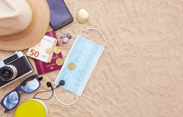 Fototapeta na wymiar Some beach items, a protective face mask, a passport and some cash, a smartphone, hat, camera and sunglasses on the beach sand. Space for text. Concept of summer in the covid-19 period.