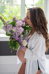 Beautiful pregnant woman in a white shirt in the bathroom near the window with a bouquet of lilac flowers. Soft selective focus.