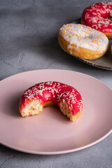 Bitten pink donut with sprinkles on pink plate near to chocolate and vanilla donuts, sweet glazed dessert food on concrete textured background, angle view macro