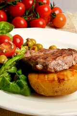 two pieces of grilled potato and grilled meat next to lettuce with tomatoes and olives on a white plate