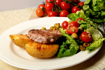 two pieces of grilled potato and grilled meat next to lettuce with tomatoes and olives on a white plate