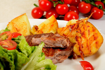 pieces of grilled steak next to potatoes grilled next to lettuce with tomatoes and olives on a white plate