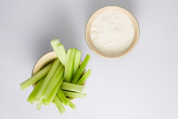 close up isolated flat lay top view shot of a bowl of crunchy juicy green celery sticks next to a white cup of blue cheese dipping sauce on a white background