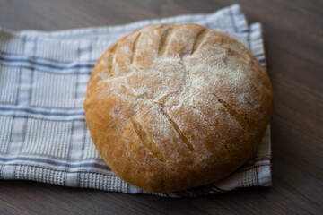 Round sourdough homemade bread with leaf scoring pattern on a wooden surface with a checked kitchen towel