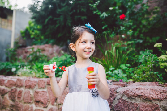 Little Caucasian girl in a white dress plays with soap bubbles in the backyard of the house on the green grass. Childhood and joy to small things. Summer and weekend. Kids toys