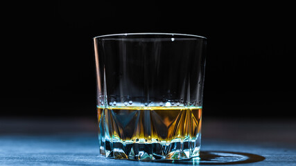 A glass of whiskey lit by a ray of light stands on a dark table and on a black background